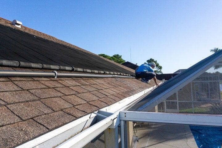Person working on a roof gutter near an adjacent structure with glass panels. Brown shingles and blue sky.