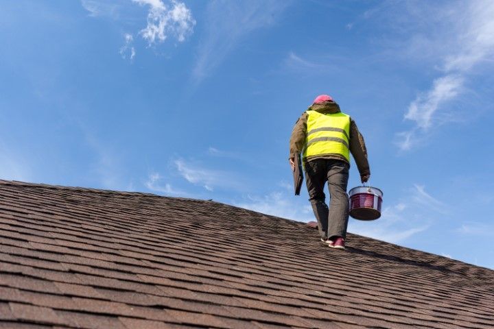 Person in a yellow vest walks on a shingled roof, carrying a paint bucket and tools, under a blue sky.