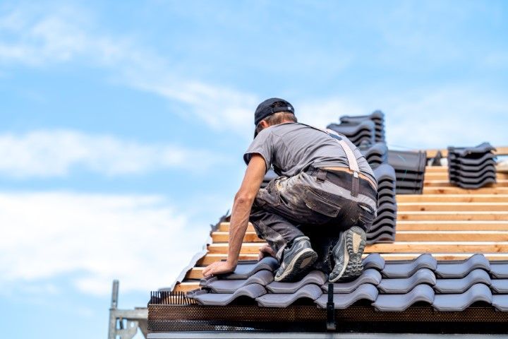 Roofer installing dark gray tiles on a wooden roof frame under a blue sky.
