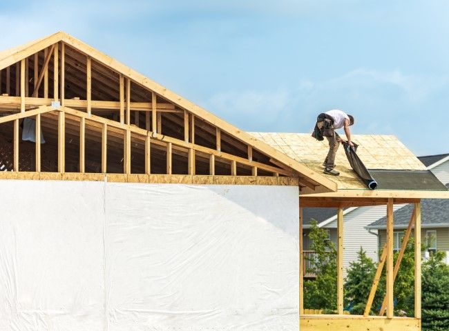 Construction worker on rooftop applying roofing material. Wooden frame house in progress with blue sky.