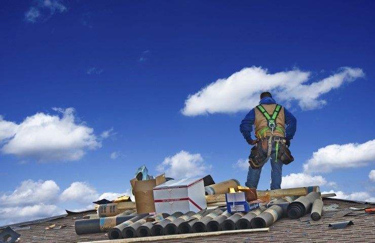Roofer wearing safety gear stands on a roof, looking out at a blue sky with clouds; tools and materials are nearby.