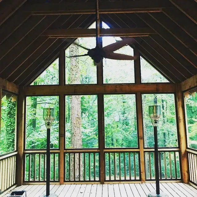 Wooden screened porch with large windows, railing, and ceiling fan overlooking lush green trees.