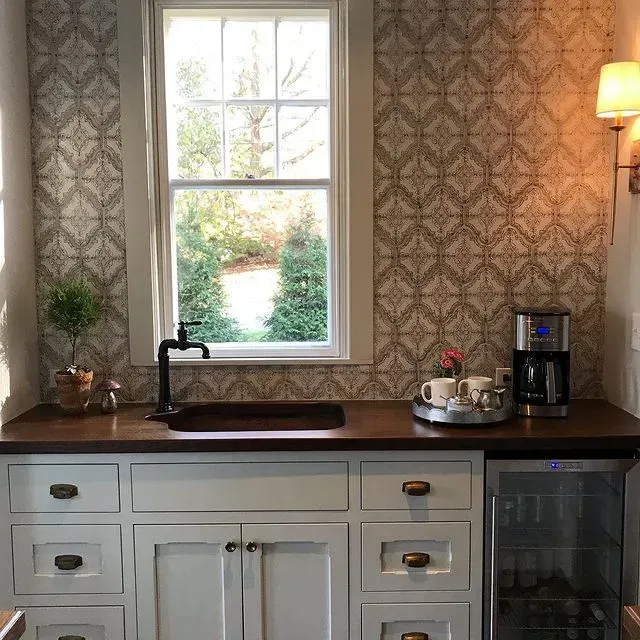 Kitchen sink under a window, with white cabinets, dark countertop, patterned wallpaper, and a coffee maker.