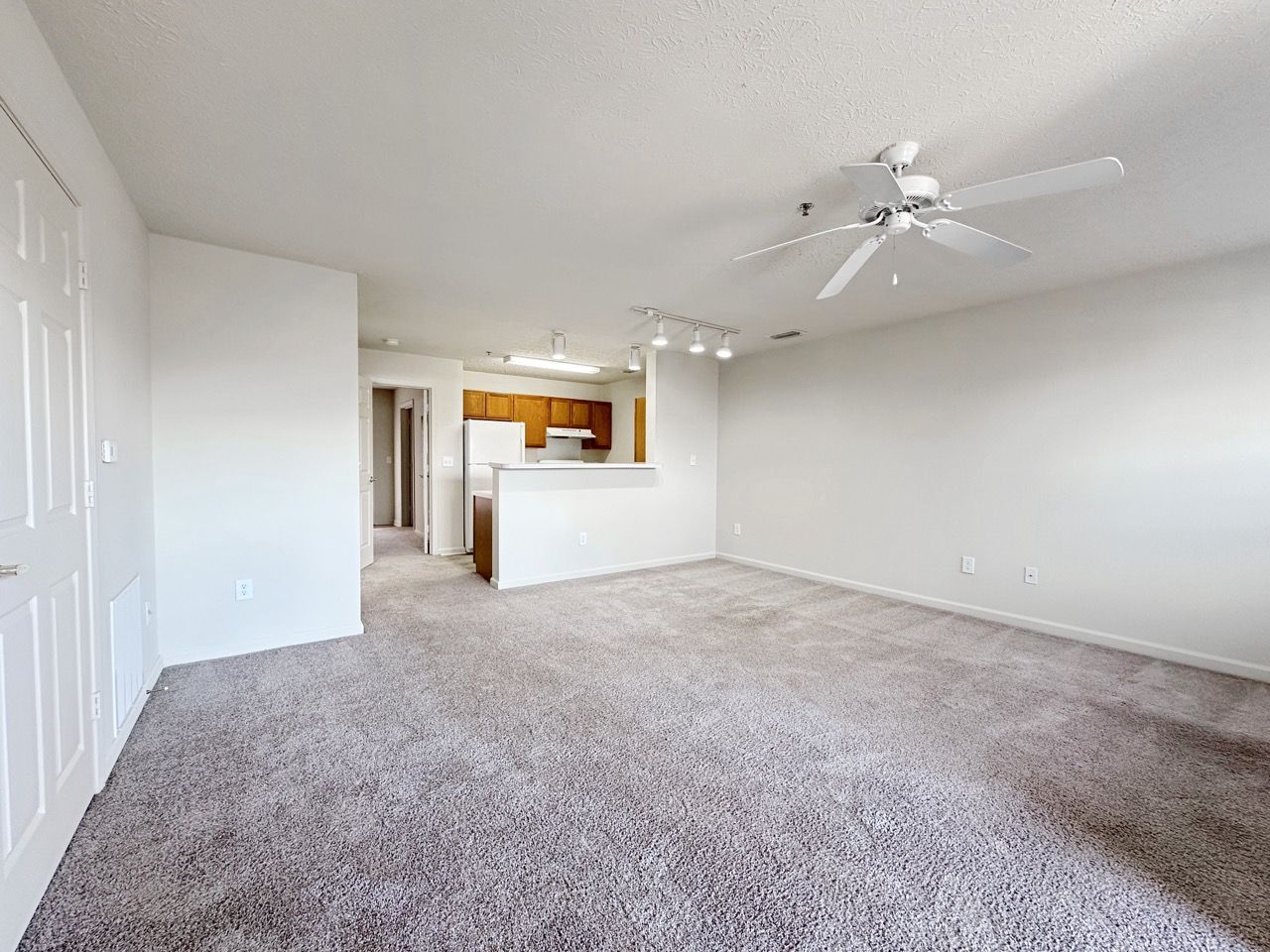 Empty living room with gray carpet, white walls, and a view into the kitchen.