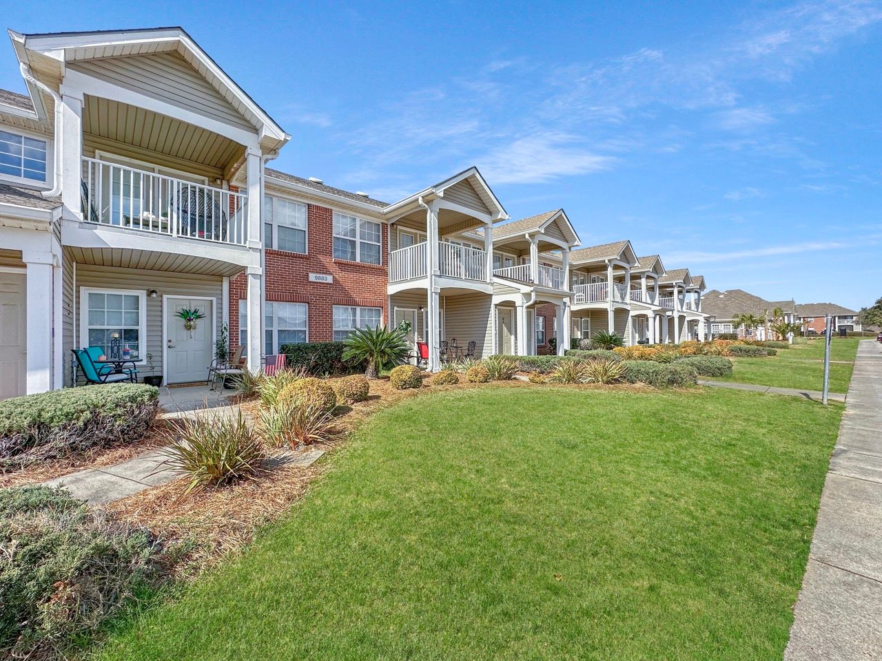 Row of two-story townhomes with white balconies, red brick and beige siding, green lawn under a blue sky.