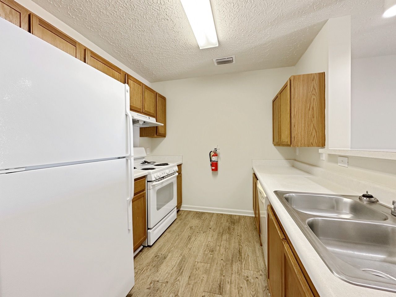 Kitchen with white appliances, wooden cabinets, and linoleum flooring. A fire extinguisher hangs on the wall.