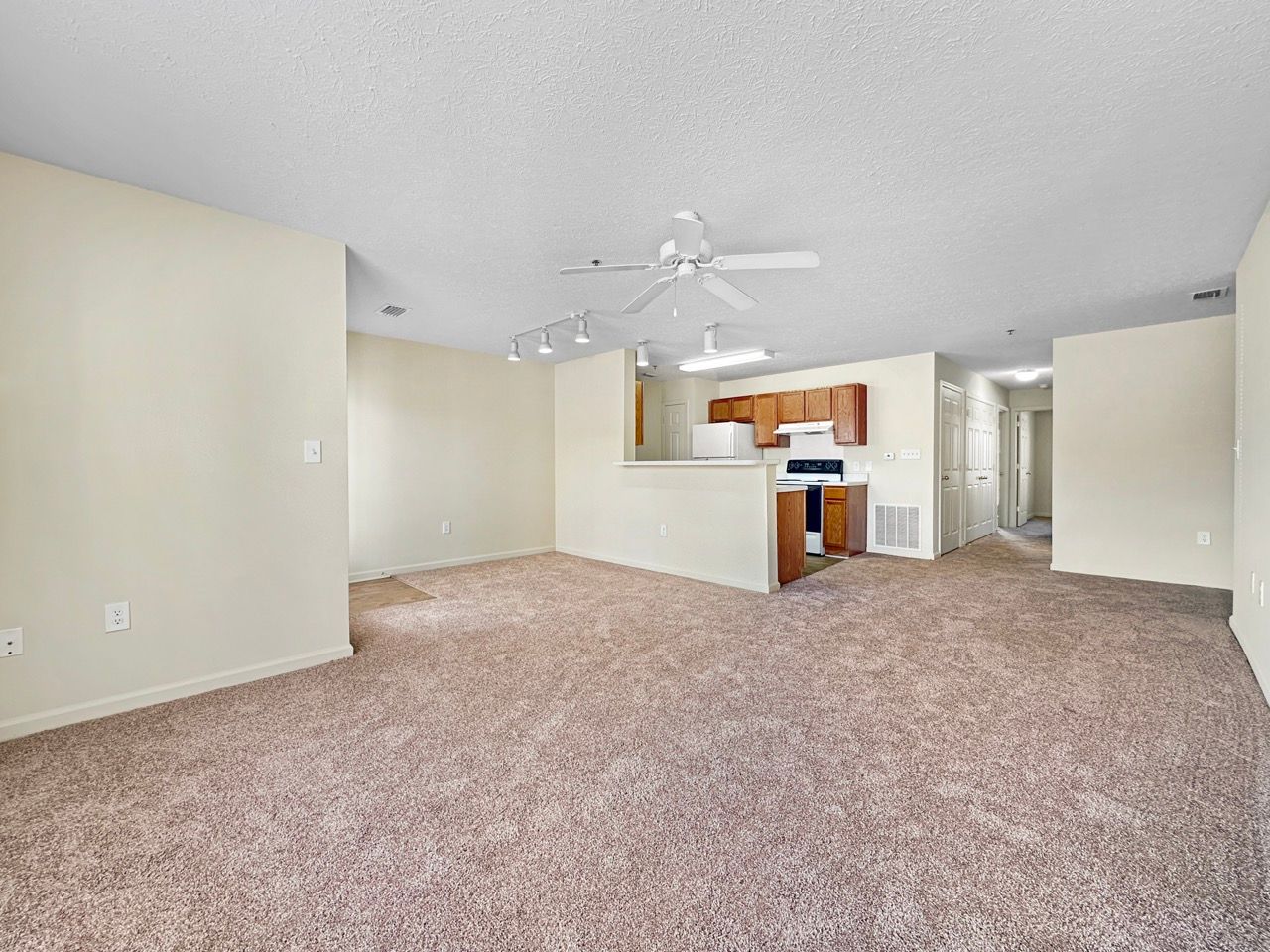 Empty living space with beige walls, carpet, and a view of the kitchen area.