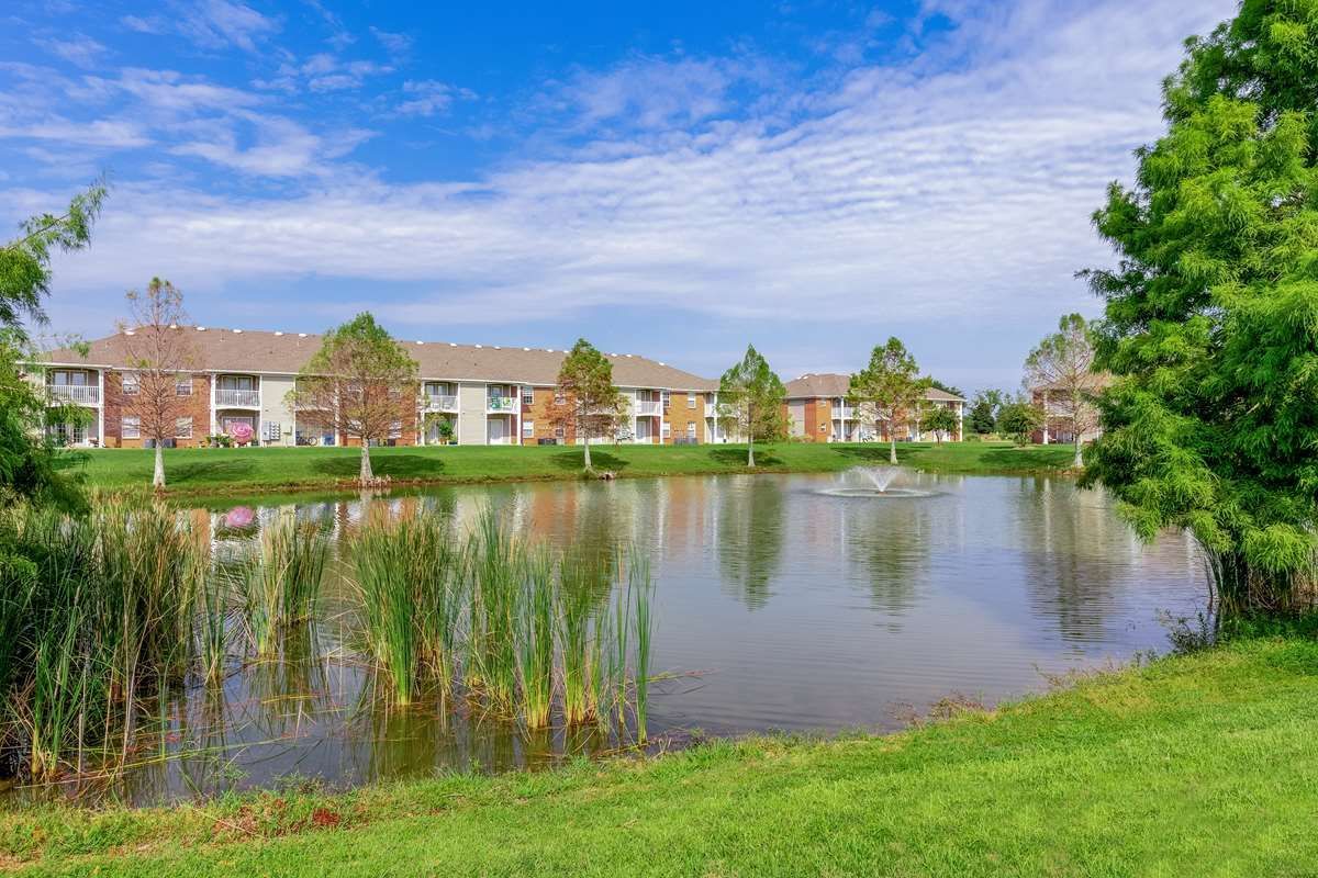 Apartment complex by a lake with fountain; green grass and trees under a blue, cloudy sky.
