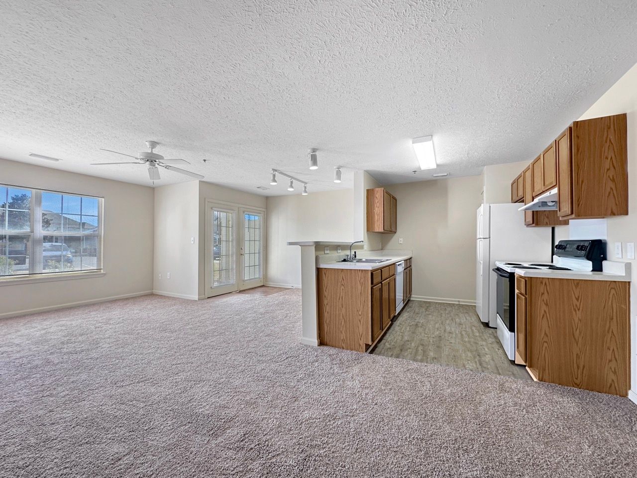 Empty apartment with a kitchen and living area, featuring light carpet and wooden cabinets.