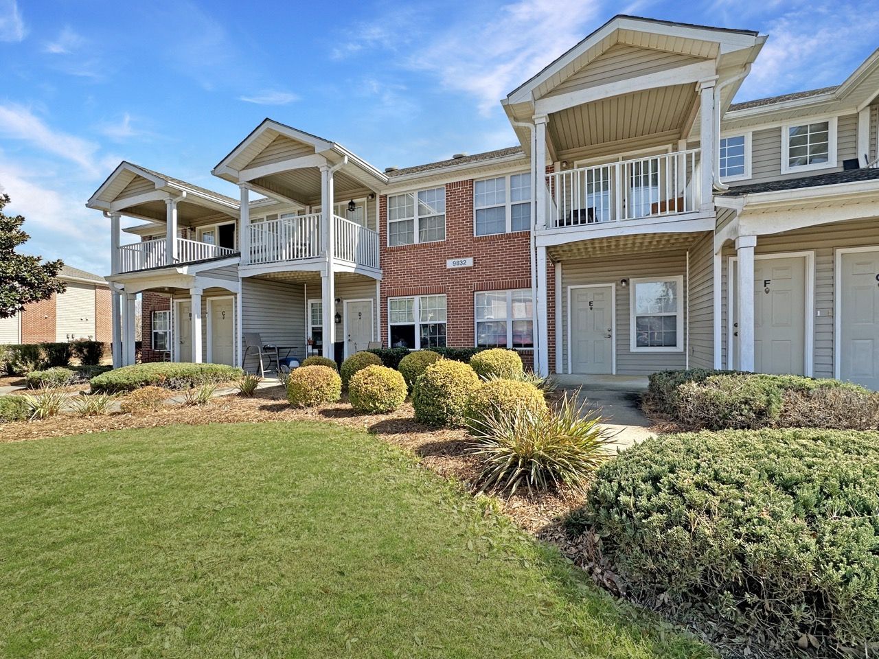 Apartment complex with beige siding, balconies, red brick accents, and green lawn under a blue sky.