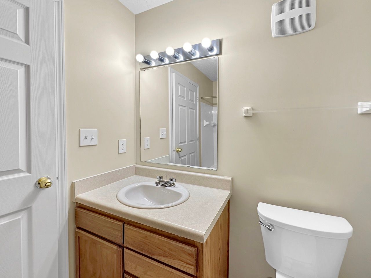 Bathroom with sink, mirror, and toilet. Beige walls, wooden cabinet, and bright lighting.