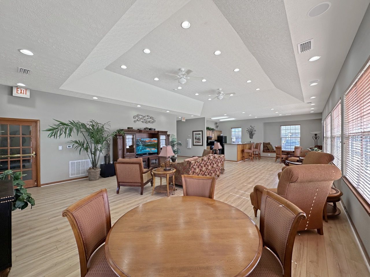 A bright, open community room with various seating areas, light wood floors, and neutral walls.