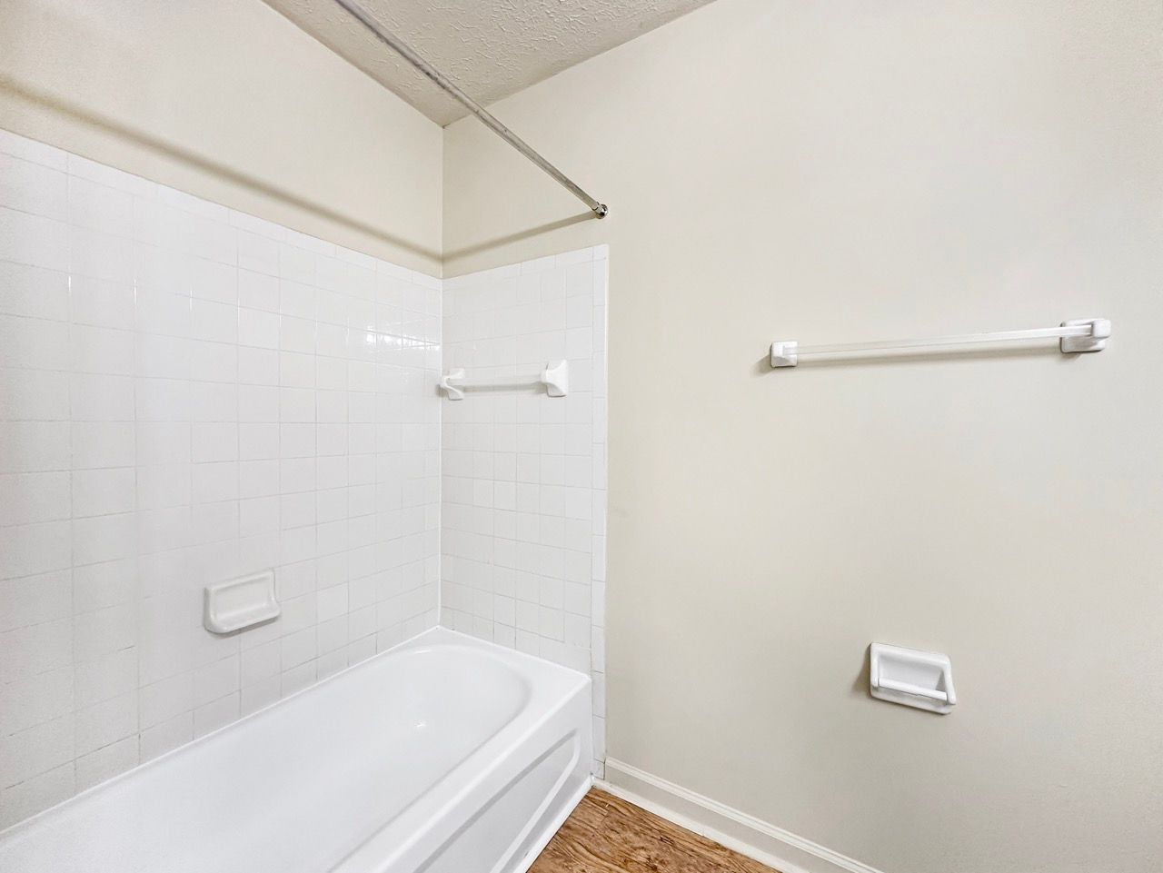 White bathroom with bathtub, towel racks, and soap dish. Light beige walls and wood-look flooring.