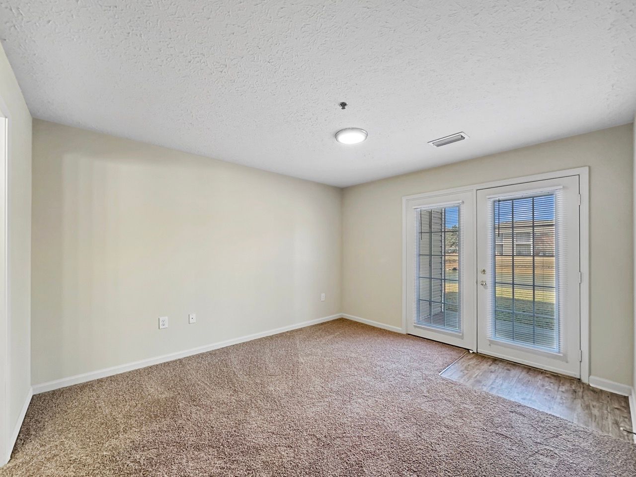 Empty room with beige walls, brown carpet, and French doors leading outside.