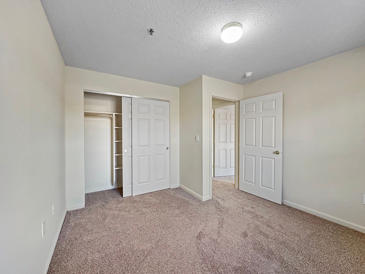 Empty bedroom with a closet, door, and doorway. Beige carpet and walls.