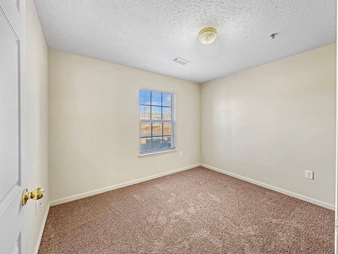 Empty room with beige walls, brown carpet, and a window; door on the left; fire alarm on the ceiling.