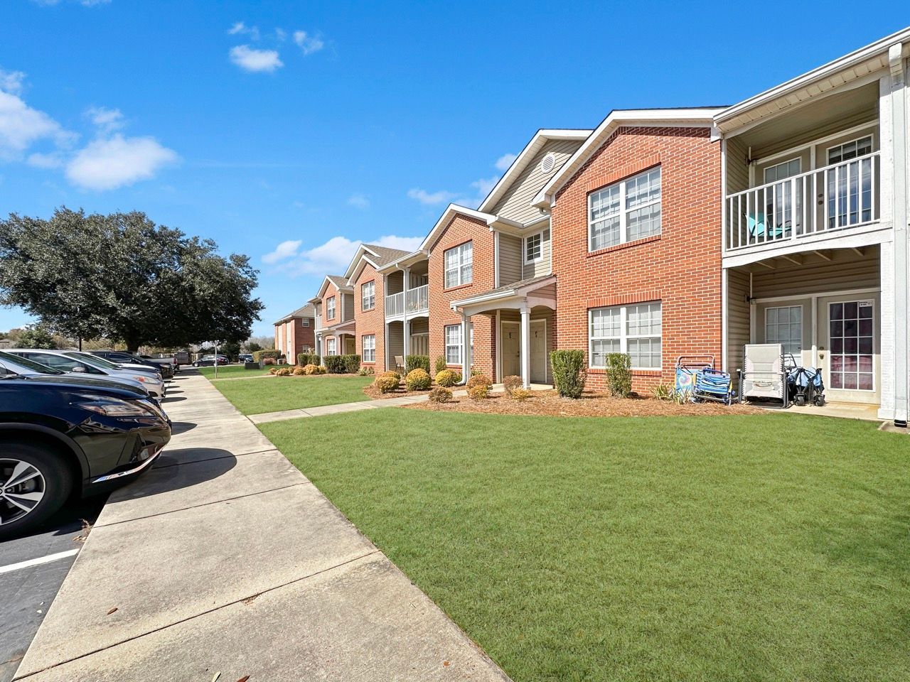 Apartment complex with brick facades, balconies, green lawn, parking lot, and blue sky.