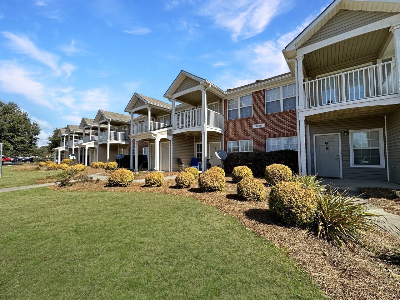 Apartment complex with multiple units, each with a balcony. Green grass and blue sky visible.