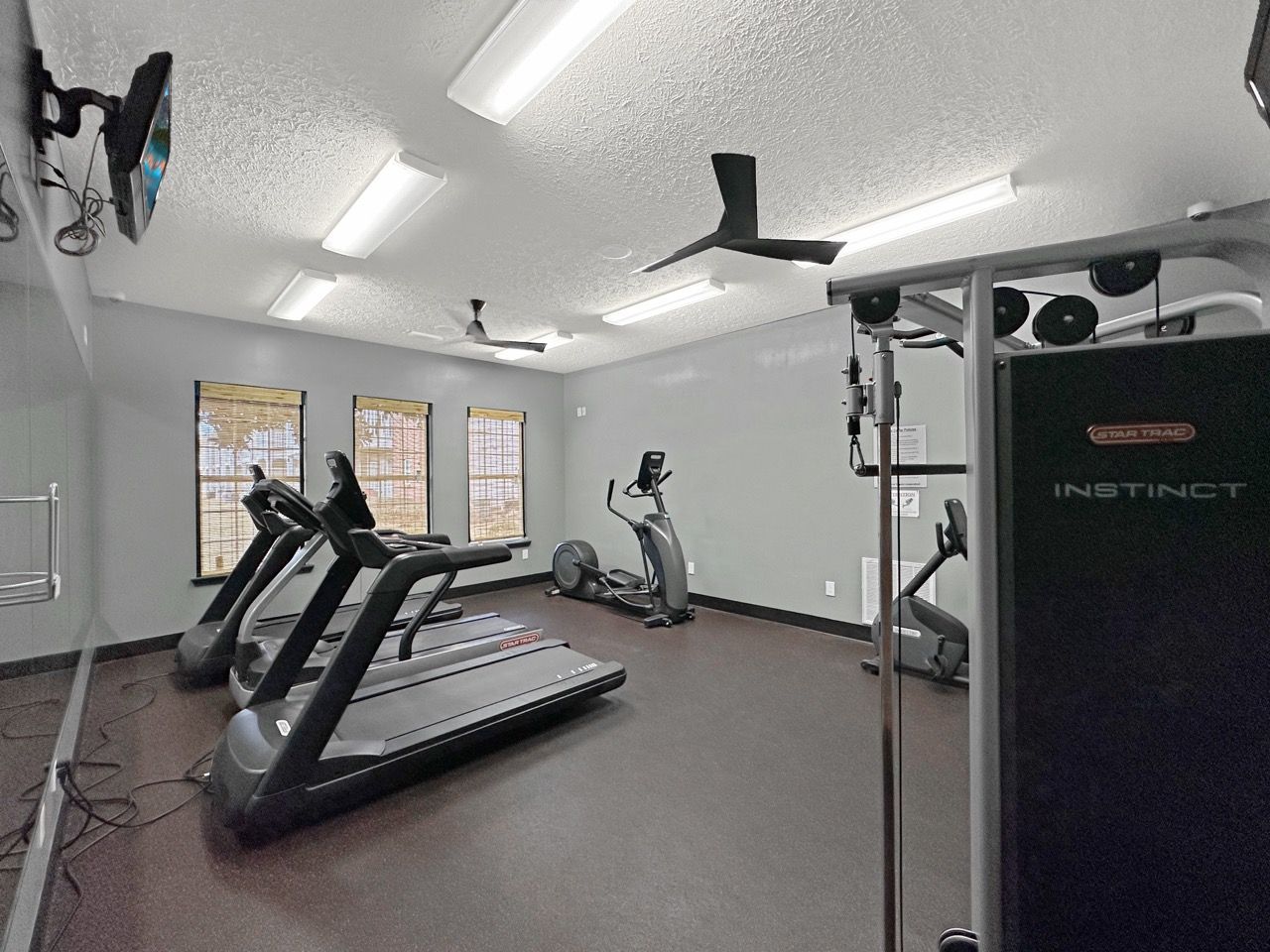 Gym with treadmills, elliptical machine, and weight machine. Gray walls, brown floor, and ceiling fans.