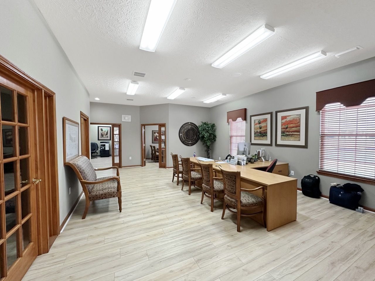 Office interior with light wood desk, chairs, artwork, and multiple doorways. Light wood flooring and grey walls.
