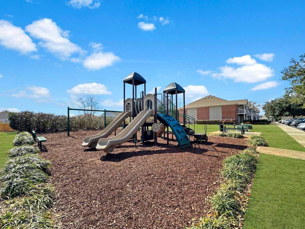 Playground with slides and climbing structure on wood chips, under a blue sky.
