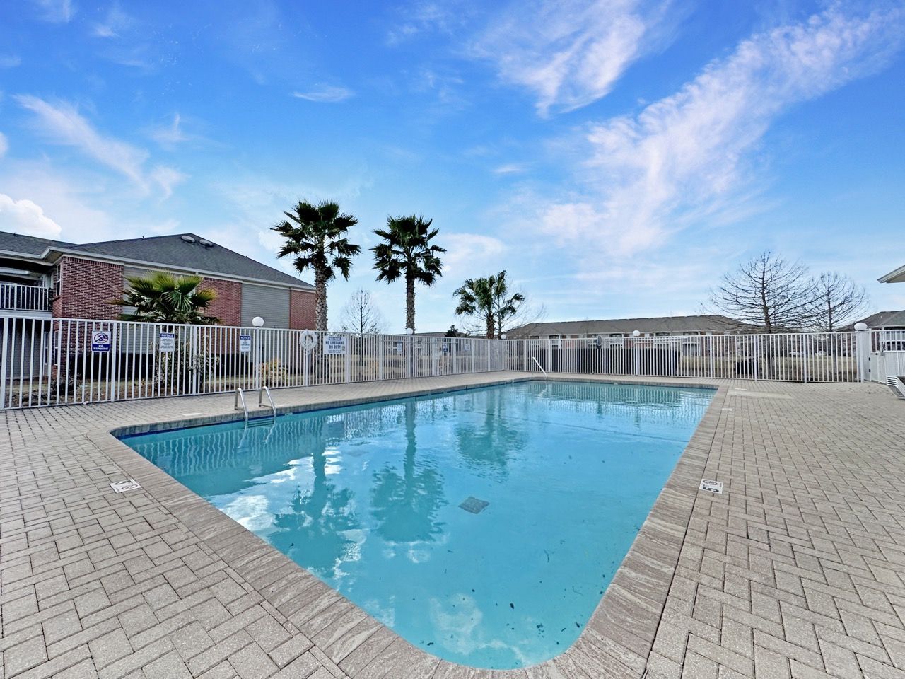 Swimming pool with brick edging, fenced, with buildings and palm trees under a blue sky.