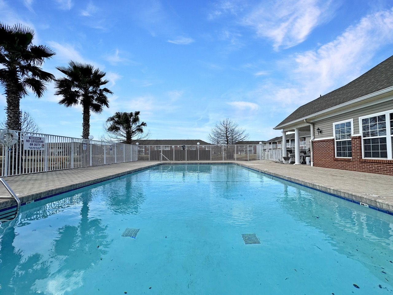 Swimming pool surrounded by a white fence, palm trees, and building under a blue sky.