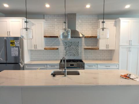 White kitchen with island, backsplash, floating shelves, stainless appliances, and hanging lights.