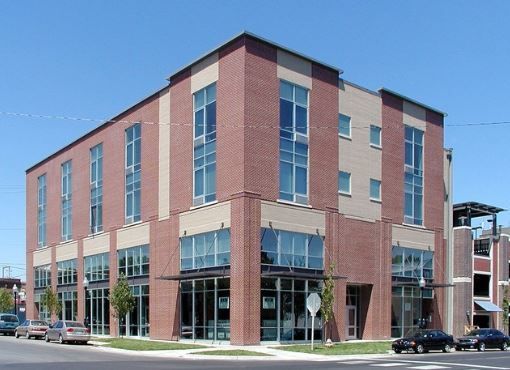 A modern three-story commercial building with red brick and tan siding on a sunny street corner with cars parked outside.