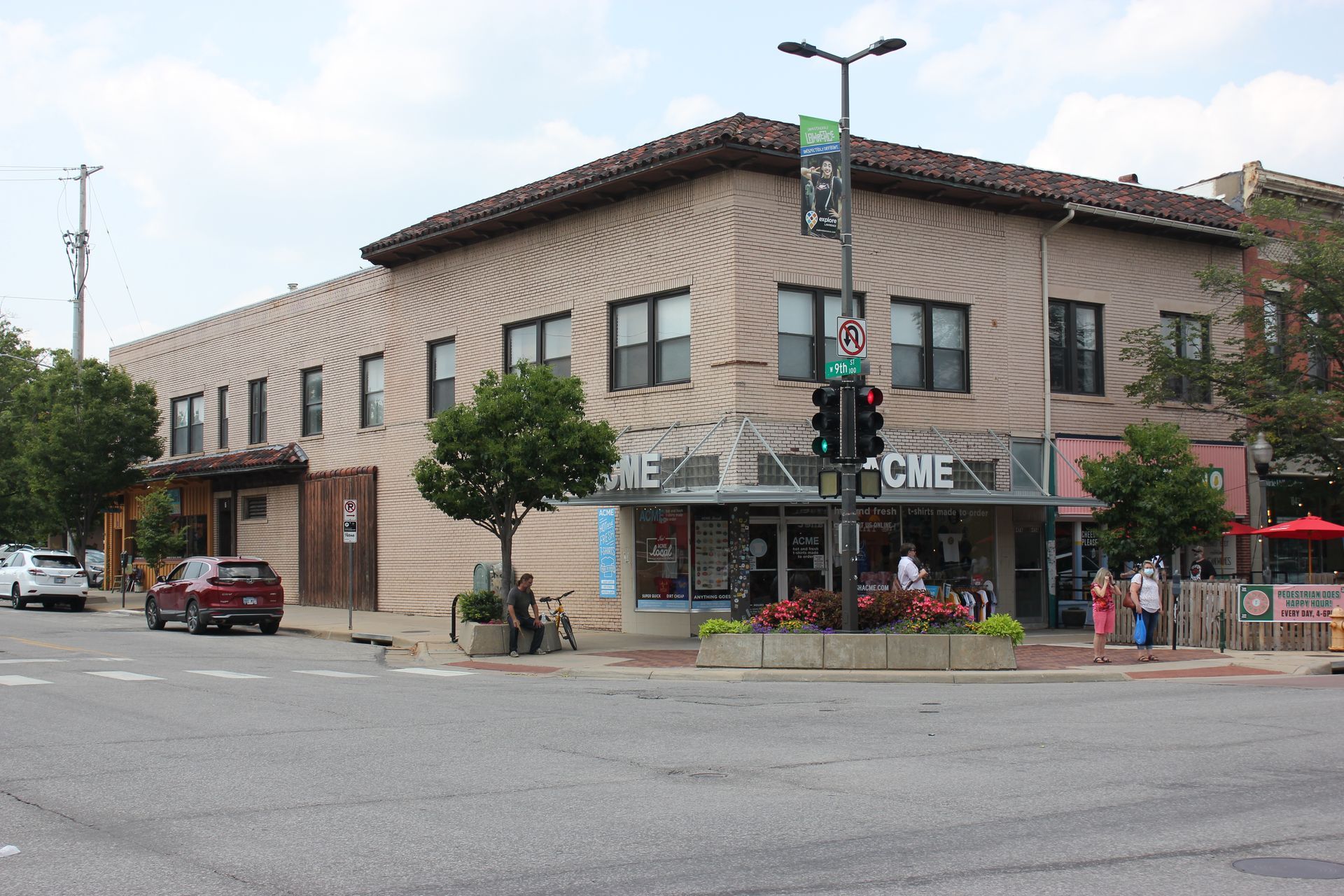 Corner building with brick facade, a cafe, and street intersection. People and parked cars visible.