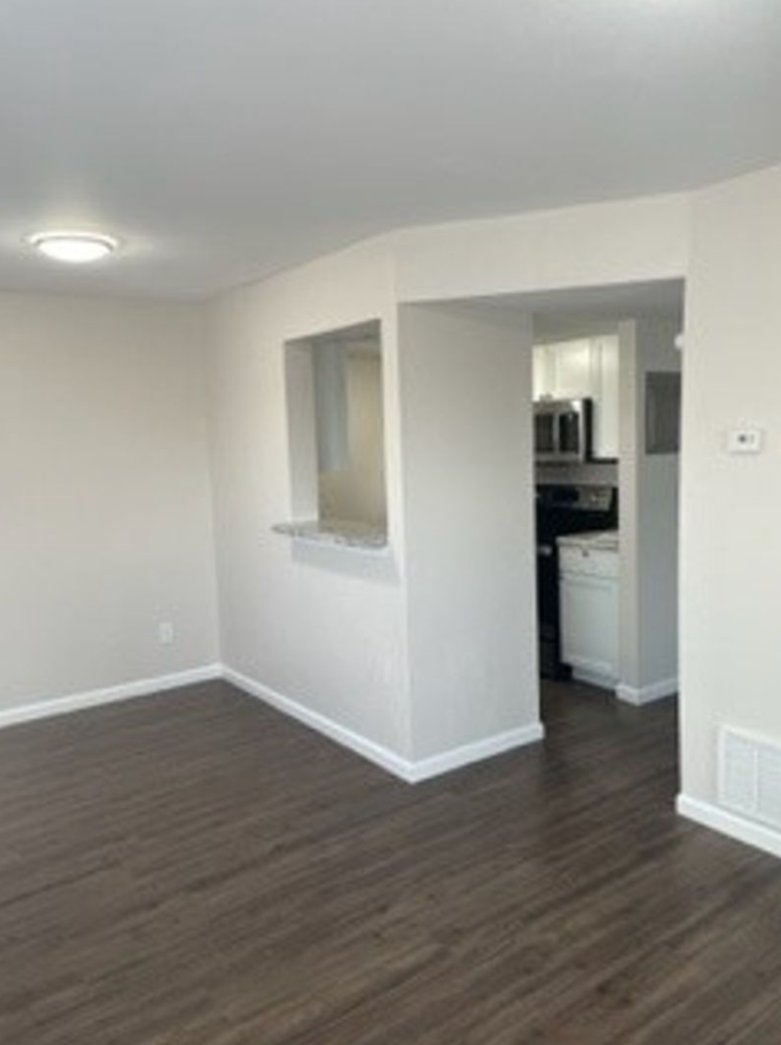 Living room with dark wood flooring, open to a white kitchen.