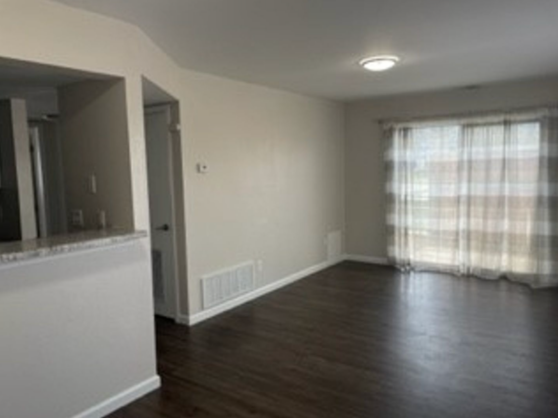 Empty living room with dark wood flooring, sliding glass door, and pale walls.