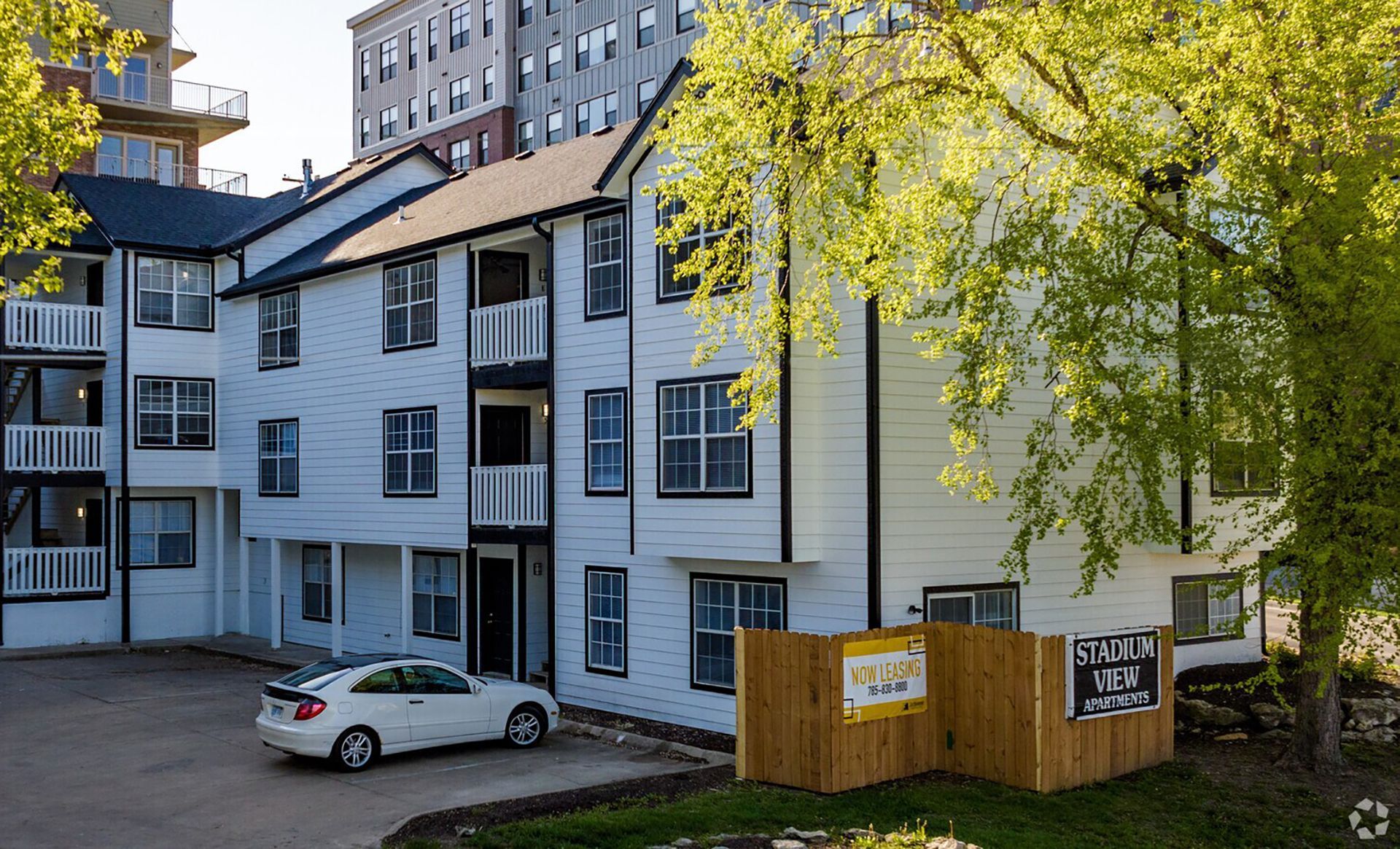 White apartment building with black trim, parked car, wooden fence, and leafy tree in front.
