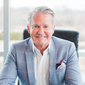 Man in a blue blazer with pocket square, sitting in an office chair, smiling.