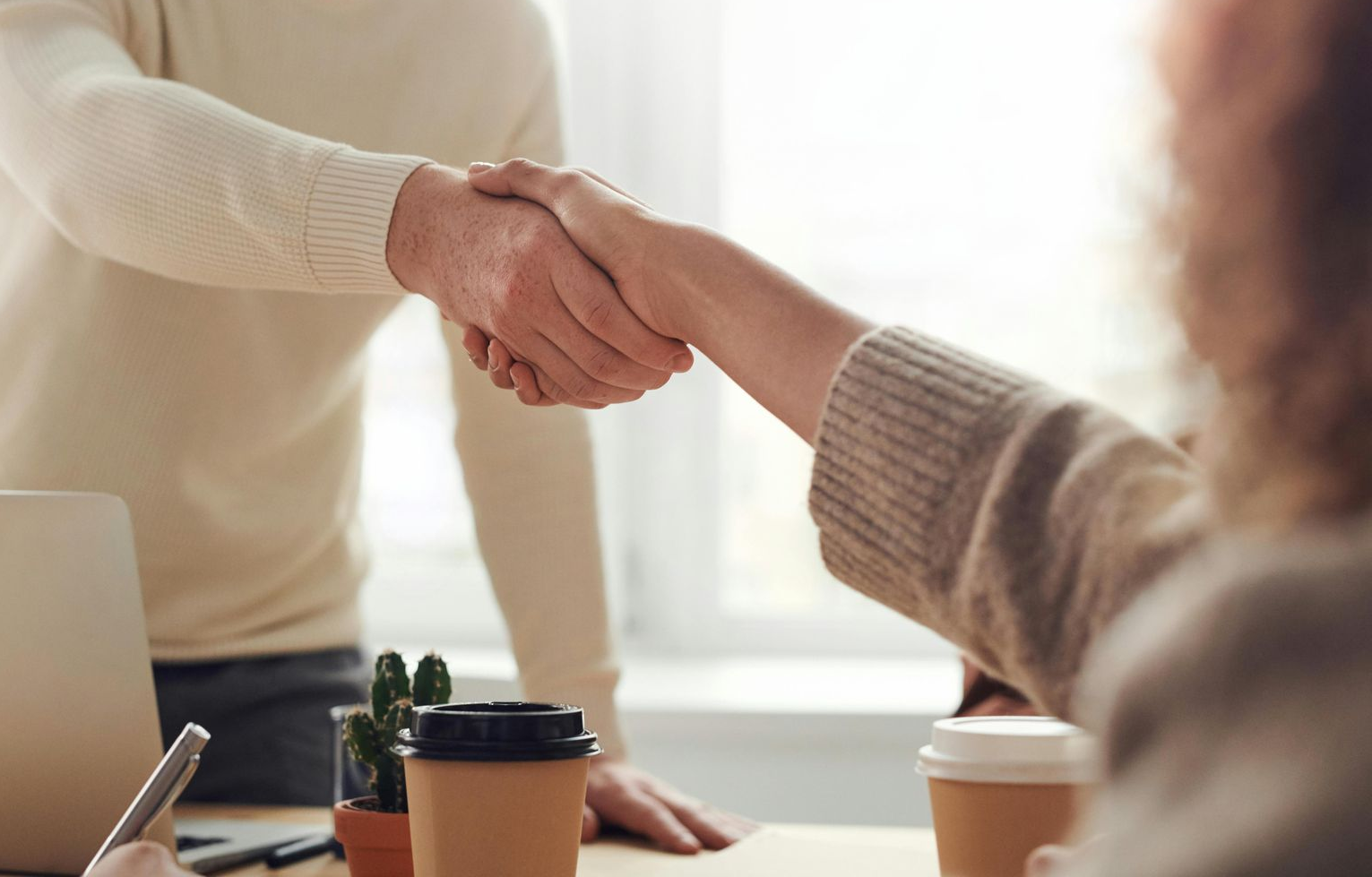 Two people shaking hands over a table with coffee cups and a laptop, possibly a business deal.