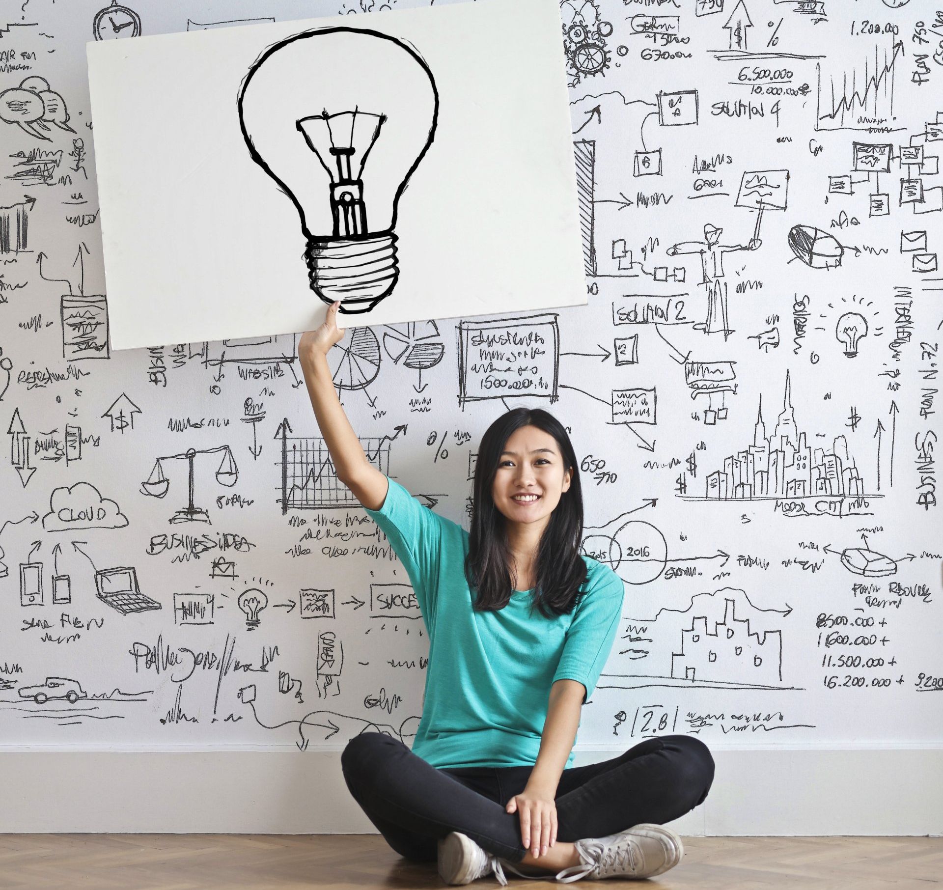 A woman is sitting on the floor holding up a sign with a light bulb drawn on it