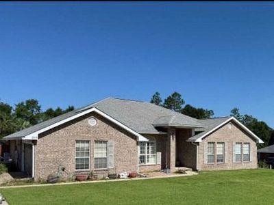 A single-story, tan brick house with a gray shingle roof sits on a green lawn under a clear blue sky.