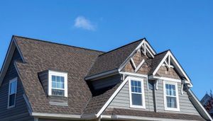 The upper story of a gray shingled house with multiple dormer windows and dark brown roof shingles against a blue sky.