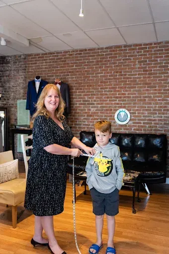 Woman measures a young boy's chest with a tape measure in a clothing store. Red brick wall in background.