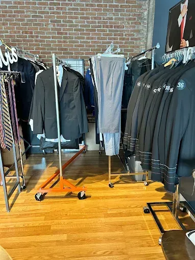 Clothing store interior with suits and ties on rolling racks against a brick wall and wood floor.