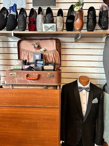 Shoes on a shelf, a vintage suitcase with accessories, and a tuxedo on a mannequin in a clothing store.