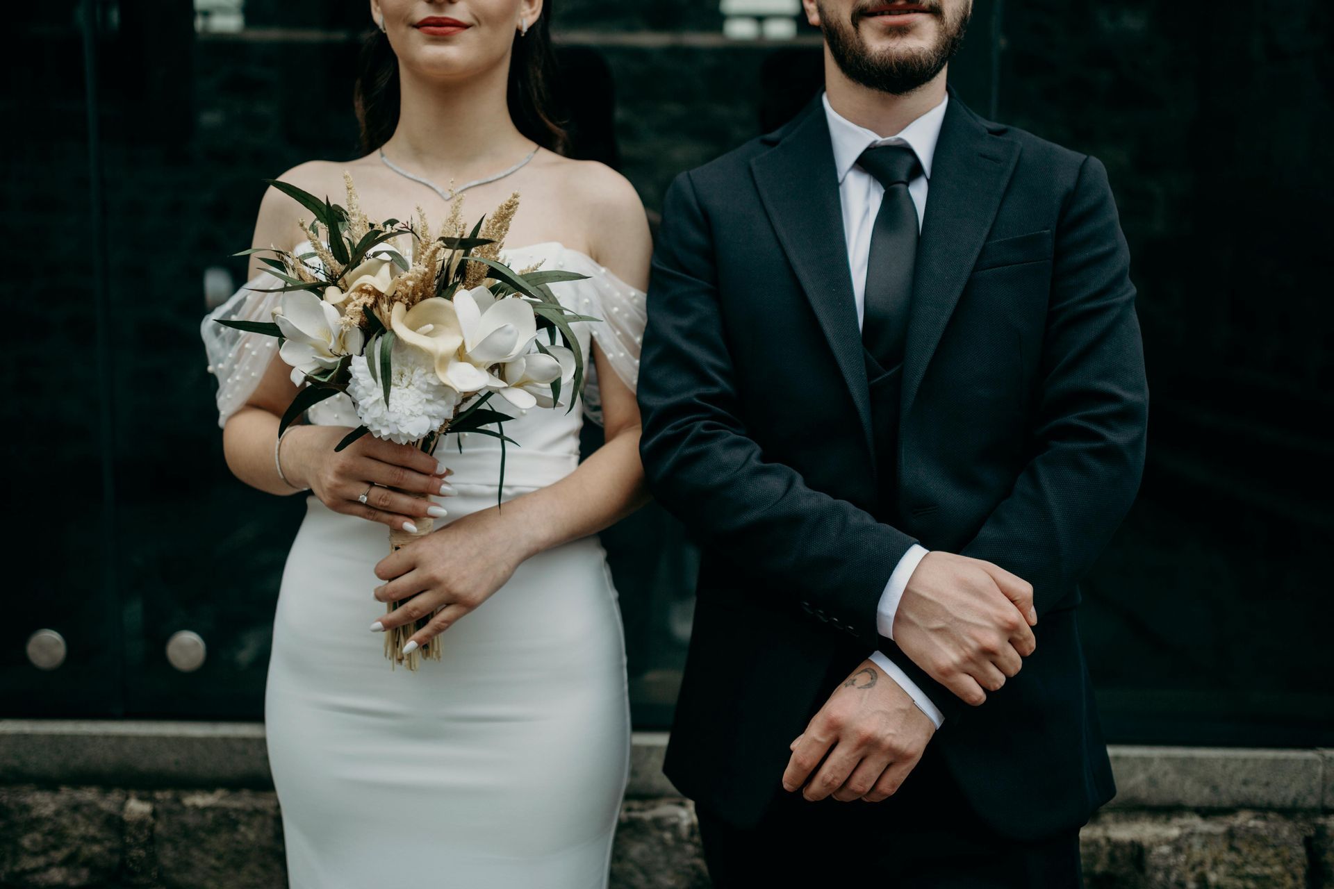 Bride and groom in formal wear, holding bouquet, standing in front of a dark background.