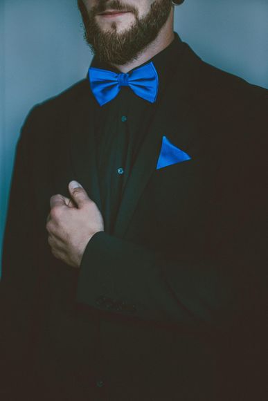 Man in black suit with blue bow tie and pocket square; hands clasped, dark background.