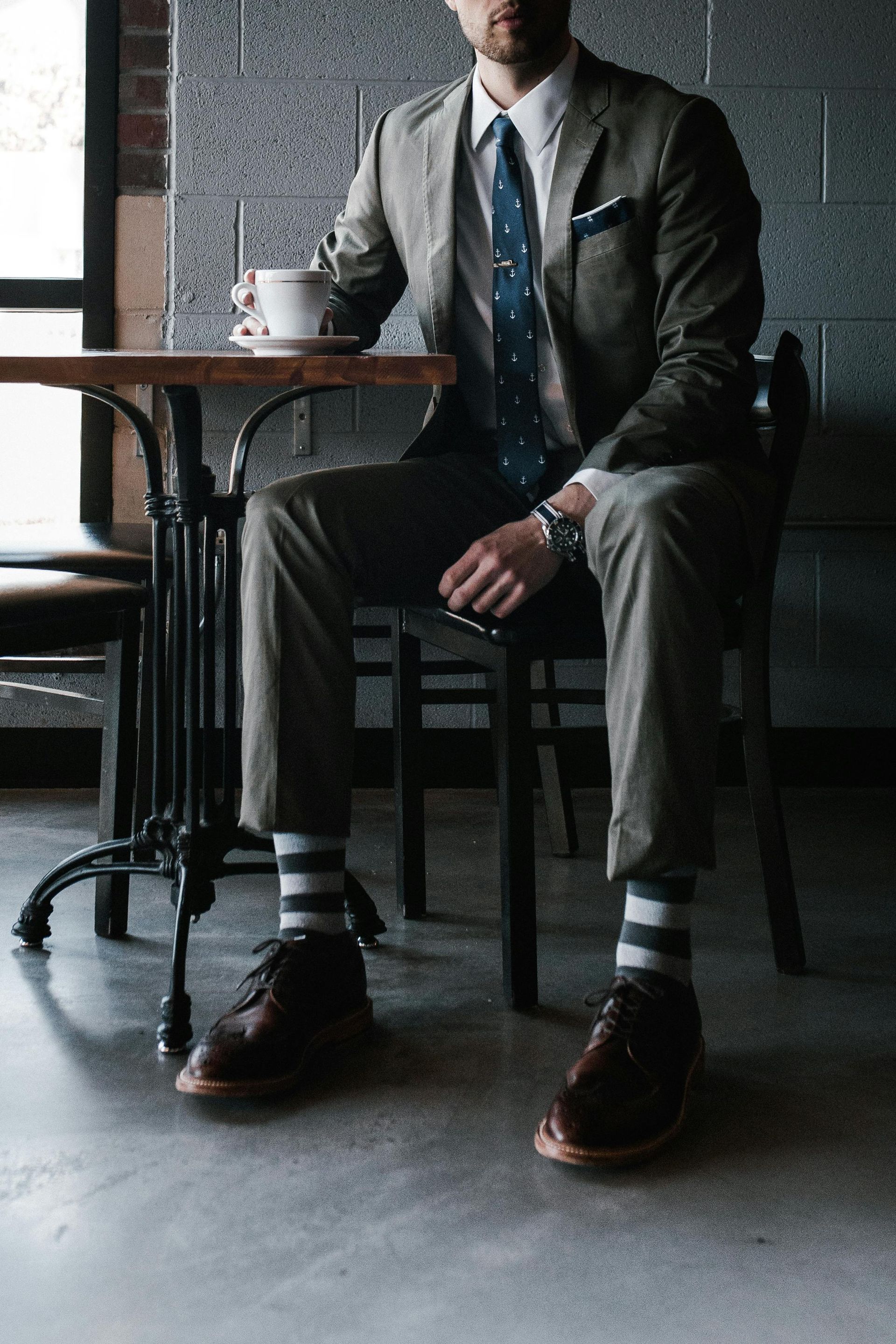 Man in suit, striped socks, and brown shoes, sitting at cafe table, holding coffee.