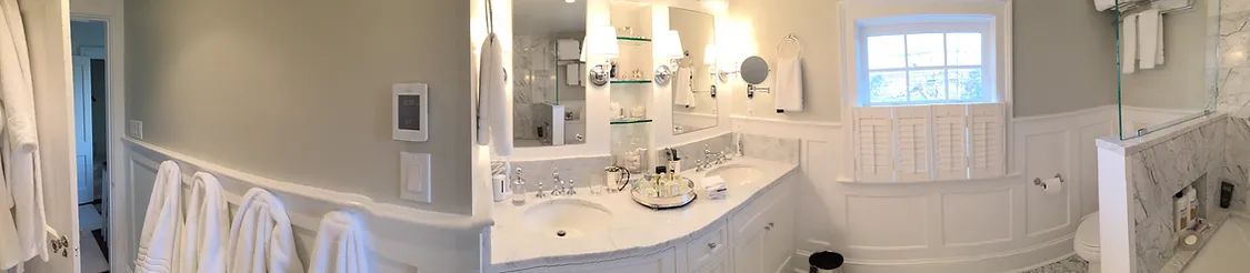 A panoramic bathroom with a double sink vanity, a window, and fluffy white robes.