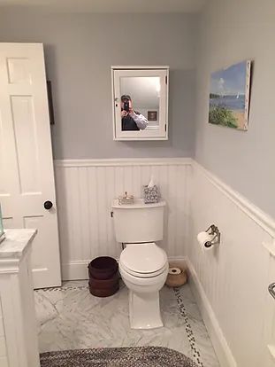 Bathroom with white wainscoting, toilet, mirror, and artwork; door on the left; blue-gray walls.