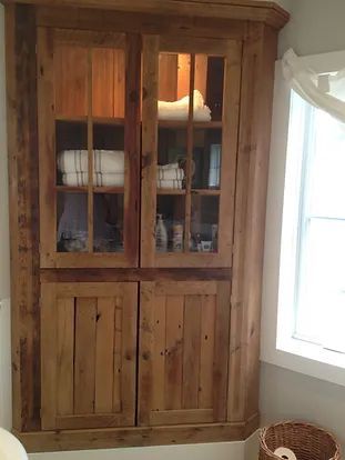 Wooden corner cabinet with glass-fronted upper doors, wooden-paneled lower doors, holding towels, toiletries, beside a window.