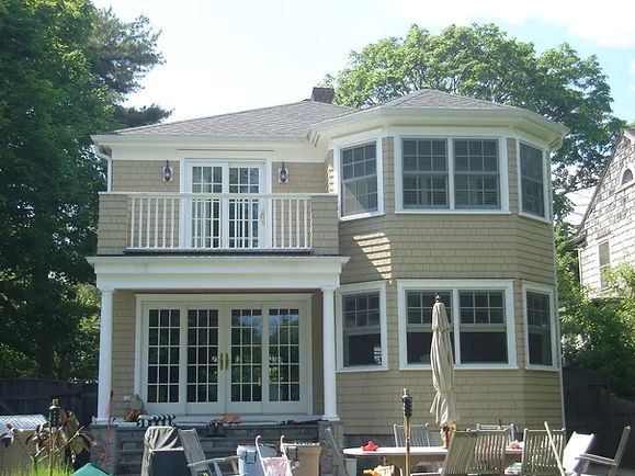 Two-story beige house with a balcony and an octagonal bay window, surrounded by trees.