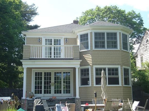 Tan house with a balcony and octagonal sunroom, surrounded by trees and an outdoor seating area.