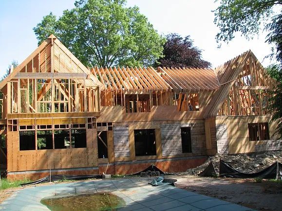 Wooden house frame under construction, with a pool in front and trees in the background.